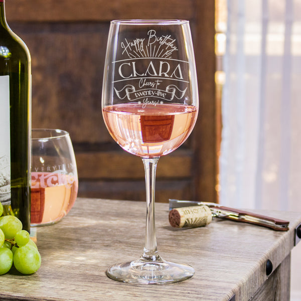 Stemmed wine glass on a table. The glass has an etched design centered on the front. The design has "Happy Birthday" in cursive rounded at the top. Below is some firework lines. Then is the name "CLARA" in between two horiztonal lines in printed all caps font. Then it says "Cheers to" in cursive, then a banner that says "twenty-five" inside, and "years!" at the bottom in cursive. The entire design is centered on top of one another.