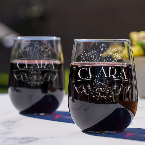 Stemless wine glass on a table. The glass has an etched design centered on the front. The design has "Happy Birthday" in cursive rounded at the top. Below is some firework lines. Then is the name "CLARA" in between two horiztonal lines in printed all caps font. Then it says "Cheers to" in cursive, then a banner that says "twenty-five" inside, and "years!" at the bottom in cursive. The entire design is centered on top of one another.