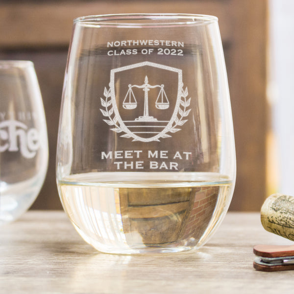 Stemless white wine glass on a table. The glass has an etched design centered. The design has "NORTHWESTERN" in all caps, and centered below says "CLASS OF 2022". Under the text is an image of a scale, inside of a shield with wreaths to represent the justice system. Below the design is the words "MEET ME AT THE BAR" in all caps centered. This design is a play on words of the bar exam and a drinking bar.
